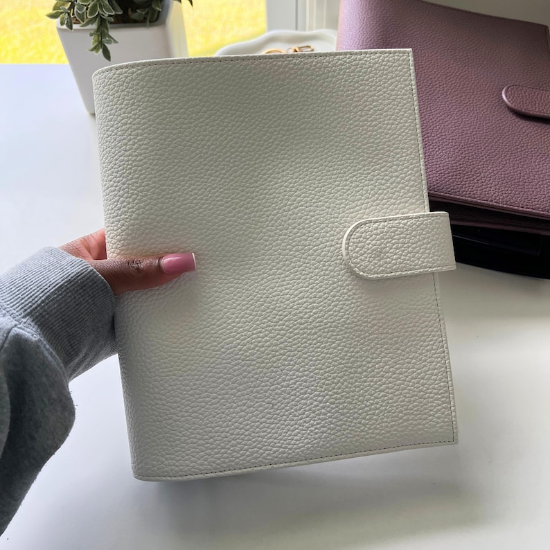 Op de afbeelding: Een witte, getextureerde leren planner met een gebogen klepsluiting. Op de achtergrond is een andere planner in mauve te zien. De planner wordt in een hand gehouden. De planner is neutraal van kleur en heeft een eenvoudig ontwerp.