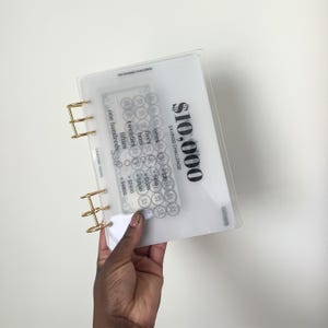 May include: A clear plastic binder with gold-toned metal rings featuring a printed $10,000 savings challenge tracker. The cover displays black text including $10,000 SAVINGS CHALLENGE, denominations like ones, fives, tens, twenties, fifties, and one hundreds.