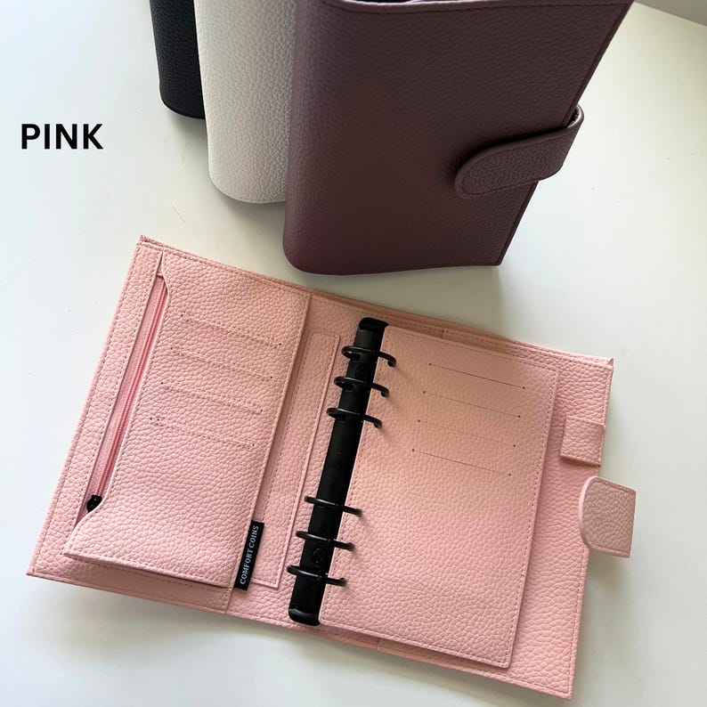 Op de afbeelding: Open roze planner met een zwarte binder en meerdere kaartsleuven. De planner is open en onthult gelinieerde pagina's en een vak met ritssluiting. Het woord "PINK" is in het zwart in de linkerbovenhoek gedrukt. Andere planners in zwart, wit en mauve zijn zichtbaar.