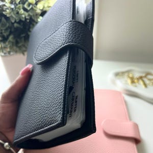 Op de afbeelding: Een zwarte leren planner met een getextureerde afwerking en een drukknoopsluiting. De planner is open en toont gelabelde verdelers voor organisatie. Een roze planner is zichtbaar op de achtergrond.