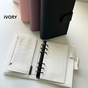 Op de afbeelding: Een open ivoorkleurige planner met een getextureerd oppervlak, met een zwart bindmechanisme en meerdere kaartsleuven. Het woord "IVORY" is in het wit gedrukt. Verschillende gesloten planners in roze, paars en zwart zijn zichtbaar op de achtergrond.