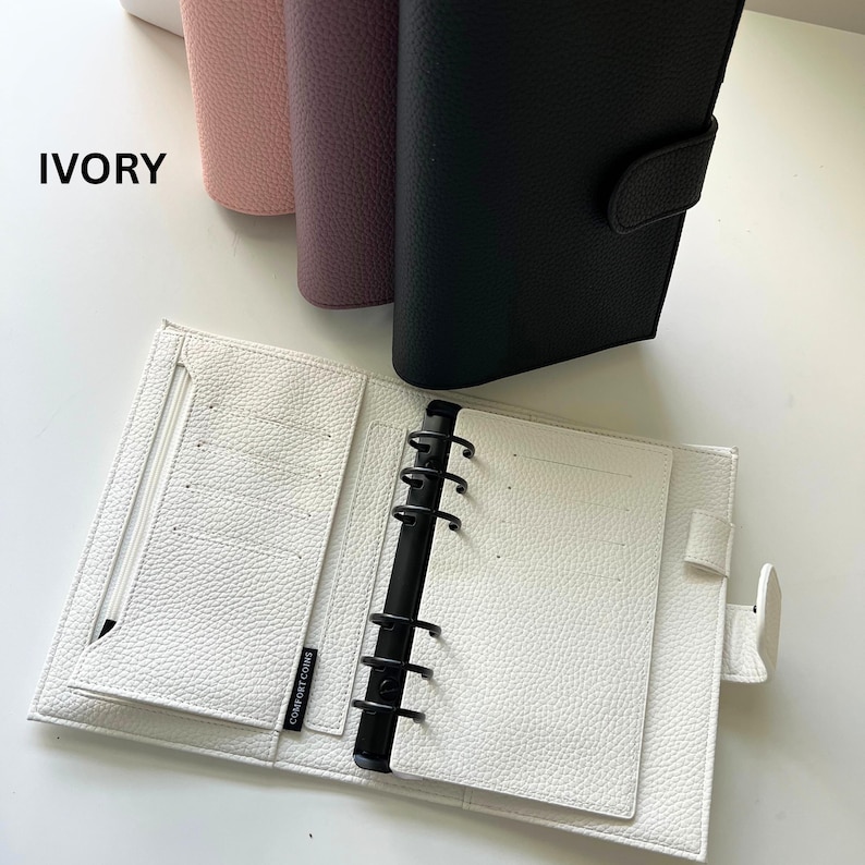 Op de afbeelding: Een open ivoorkleurige planner met een getextureerd oppervlak, met een zwart bindmechanisme en meerdere kaartsleuven. Het woord "IVORY" is in het wit gedrukt. Verschillende gesloten planners in roze, paars en zwart zijn zichtbaar op de achtergrond.
