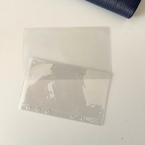 Op de afbeelding: Transparante plastic insteekhoes met een rij versterkte gaten langs de onderrand. De hoes is aan de bovenkant open en heeft een vak aan de voorkant. Op de achtergrond is een donkerblauw leren item zichtbaar.