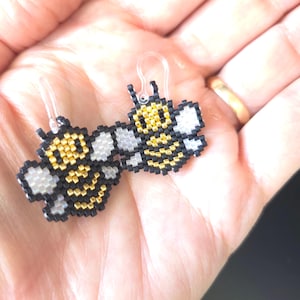 Könnte beinhalten: Ein Paar Perlen-Ohrringe mit Bienenmotiv. Die Ohrringe haben einen gelb-schwarzen Körper mit weißen Flügeln. Sie sind an durchsichtigen Kunststoffhaken befestigt.