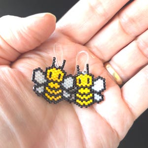 Könnte beinhalten: Ein Paar handgefertigte Ohrringe in Form von Bienen. Die Ohrringe bestehen aus gelben, schwarzen und weißen Perlen und haben transparente Kunststoffhaken. Ein verspieltes und charmantes Accessoire.