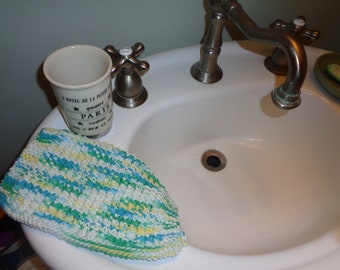 May include: A white ceramic mug with black text that reads "L'hotel de la Petite quatre Paris" and a green, blue, and yellow striped knitted washcloth in a white porcelain sink with a silver faucet.