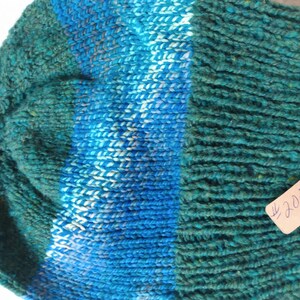 May include: A green and blue knitted beanie with a white stripe. The beanie has a tag attached that says "#205".