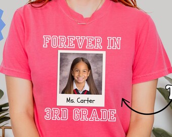 May include: A pink t-shirt with the words "FOREVER IN" and "3RD GRADE" printed on it. A photo of a student is in the center, with the teacher's name below. The shirt also has the text "More Colors Available".