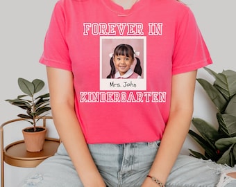 May include: Pink t-shirt with the text "FOREVER IN KINDERGARTEN" in white. A framed photo of a young girl with the text "Mrs. John" is in the center of the shirt.