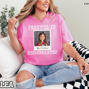 May include: A pink t-shirt with the words "FOREVER IN KINDERGARTEN" in white block letters. A photo of a young girl is in the center, with the name "Ms. Froman" below. The shirt is worn with blue jeans.