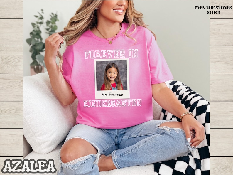 May include: A pink t-shirt with the words "FOREVER IN KINDERGARTEN" in white block letters. A photo of a young girl is in the center, with the name "Ms. Froman" below. The shirt is worn with blue jeans.