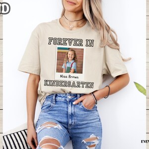 May include: Sand-colored t-shirt with the text "FOREVER IN KINDERGARTEN" in a black, blocky font. A photo of a young girl is centered within the text. The shirt is worn by a person wearing blue jeans.