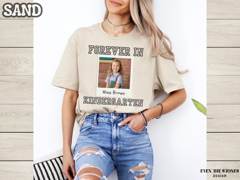 May include: Sand-colored t-shirt with the text "FOREVER IN KINDERGARTEN" in a black, blocky font. A photo of a young girl is centered within the text. The shirt is worn by a person wearing blue jeans.