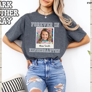 May include: Dark heather gray t-shirt with the words "FOREVER IN KINDERGARTEN" and a photo of a young girl. The text "Miss Smith" is below the photo. The shirt is worn by a person wearing blue jeans.