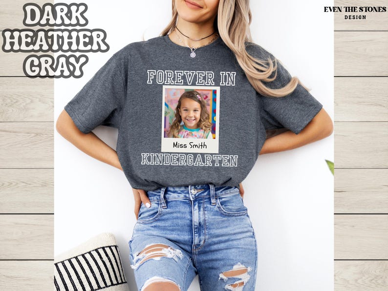 May include: Dark heather gray t-shirt with the words "FOREVER IN KINDERGARTEN" and a photo of a young girl. The text "Miss Smith" is below the photo. The shirt is worn by a person wearing blue jeans.