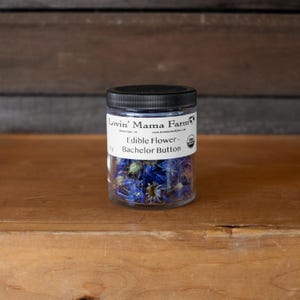 May include: A clear jar with a black lid filled with blue edible Bachelor Button flowers. The jar has a white label with the text "Lovin' Mama Farm" and "Edible Flower - Bachelor Button". The jar is sitting on a wooden surface.