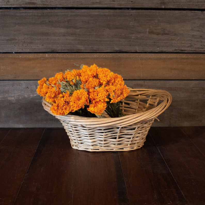 K&ouml;nnte beinhalten: Ein geflochtener Weidenkorb gef&uuml;llt mit leuchtend orangefarbenen Ringelblumen. Der Korb hat einen gebogenen Griff und steht auf einer dunklen Holzoberfl&auml;che und einem Hintergrund aus Holzplanken. Die Blumen sind in voller Bl&uuml;te.