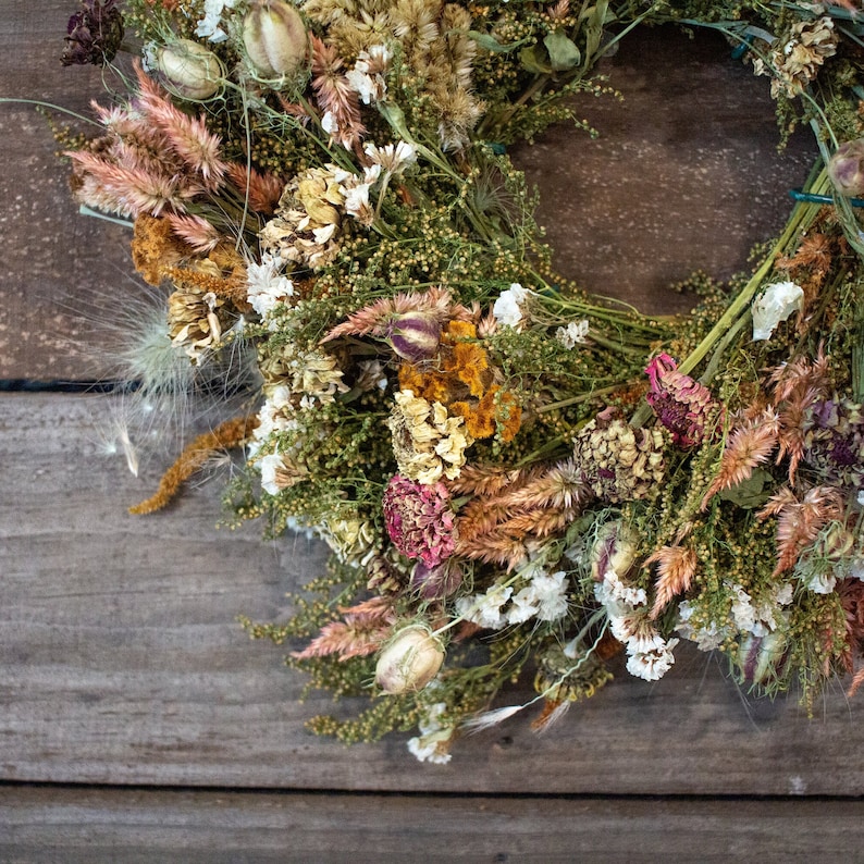 May include: A dried flower wreath with a variety of textures and colors. The wreath features dried flowers in shades of pink, white, yellow, and brown, arranged in a circular shape. The background is a rustic wooden surface.