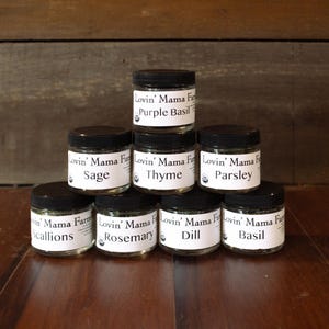 May include: A pyramid of eight small glass jars with black lids, each containing dried herbs. The labels on the jars read "Lovin' Mama Farm" and the name of the herb, including Sage, Thyme, Parsley, Scallions, Rosemary, Dill, Basil, and Purple Basil.