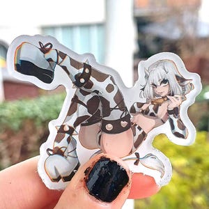 Peut inclure: Un autocollant transparent représentant une cowgirl de dessin animé aux cheveux blancs et une tenue à imprimé vache. L'autocollant a une bordure blanche et est tenu par une main avec du vernis à ongles noir. La cowgirl est dans une pose suggestive.