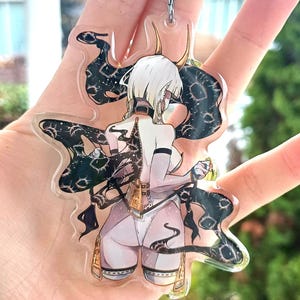 Könnte beinhalten: Ein klarer Acryl-Schlüsselanhänger mit einem stilisierten Anime-Charakter mit weißen Haaren, Hörnern und einem freizügigen Outfit. Der Charakter ist von schwarzen, wirbelnden, gemusterten Elementen umgeben. Der Schlüsselanhänger hat einen silbernen Metallring.