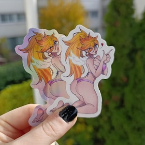 Könnte beinhalten: Zwei holografische Aufkleber mit einer Cartoon-Illustration einer Frau mit langen, orangefarbenen Haaren, die einen rosa Bikini trägt. Die Frau kniet und zeigt mit einem Herzen über ihrem Finger. Die Aufkleber haben ein glänzendes, irisierendes Finish.