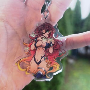 May include: A clear acrylic keychain featuring a cartoon illustration of a woman with long red hair and a blue bikini. The woman is making a heart shape with her hands. The keychain is attached to a silver chain.
