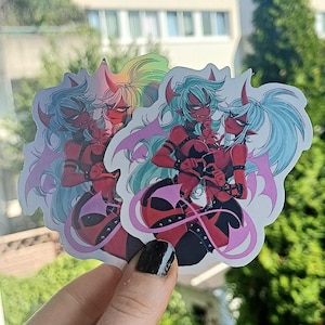 May include: A stack of stickers depicting anime-style characters with red skin, horns, and devil tails. The characters have light blue and rainbow hair, wearing black and red outfits with pink bat wings. The stickers are held in front of a window.
