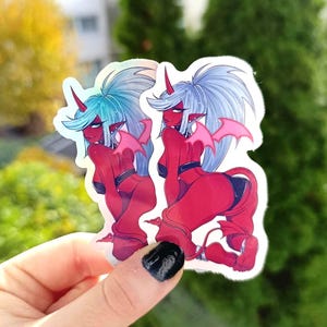 Könnte beinhalten: Zwei holografische Aufkleber mit einem rothäutigen Anime-Charakter mit blauen oder weißen Haaren, roten Hörnern und rosa Fledermausflügeln. Die Figur kniet und trägt schwarze Unterwäsche und ein passendes Oberteil. Die Aufkleber werden vor grünem Hintergrund gehalten.