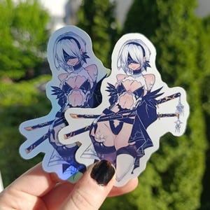 May include: Two holographic stickers featuring a character with white hair, a blindfold, and a black and white outfit. The character is holding two swords. The stickers are held in a hand against a blurred green background.