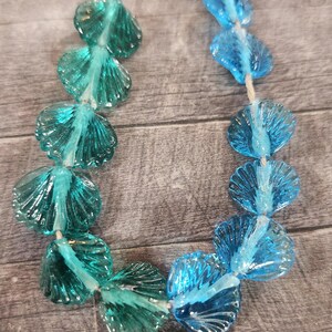 May include: A necklace featuring translucent, shell-shaped beads in shades of teal and blue. The beads are strung together with a thin, off-white cord, creating a delicate and ocean-inspired accessory.