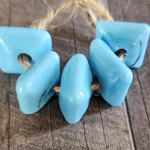 May include: Five light blue glass beads with a smooth, glossy finish and rounded square shapes. The beads are strung together on a piece of natural tan twine, showing subtle dark swirls within the opaque light blue glass material.