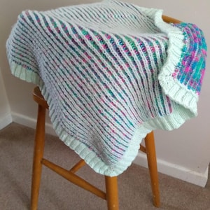 Puede incluir: Una manta de bebé de punto a rayas azul claro y blanco con un borde colorido. La manta está drapeada sobre una silla de madera.