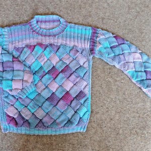 Könnte beinhalten: Ein gestrickter Pullover mit einem bunten Patchwork-Muster in Blautönen, Lila und Rosa. Der Pullover hat einen Rundhalsausschnitt und lange Ärmel.