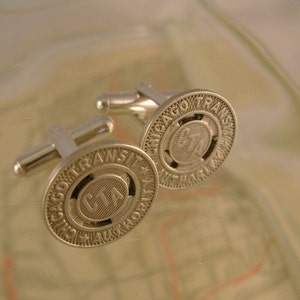 May include: A pair of silver cufflinks featuring the Chicago Transit Authority (CTA) logo. The cufflinks are circular and have a raised design with the words "Chicago Transit Authority" and "CTA" in the centre.