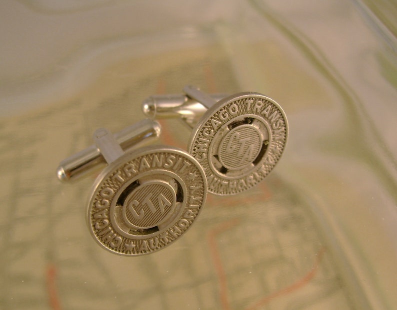 May include: A pair of silver cufflinks featuring the Chicago Transit Authority (CTA) logo. The cufflinks are circular and have a raised design with the words "Chicago Transit Authority" and "CTA" in the centre.