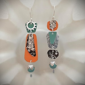 May include: A pair of mismatched dangle earrings featuring silver-tone hooks. The left earring has a teal gear, an orange trapezoid, and a black and white patterned piece. The right earring has an orange hexagon, a teal triangle, a silver embossed piece, and a black swirl patterned piece. Both end with a teal bead.