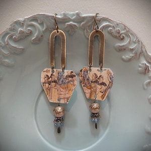 May include: A pair of dangle earrings featuring an inverted U-shaped brass component attached to a flat, irregular pentagonal charm with a printed historical illustration. Each earring ends with a metallic bead, a decorative cap, and a small blue crystal.