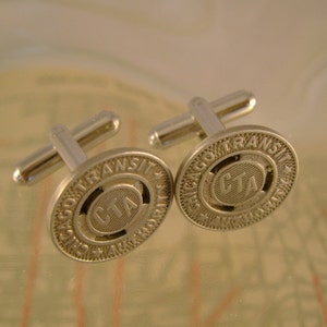 May include: A pair of silver cufflinks featuring the Chicago Transit Authority (CTA) logo. The cufflinks are round and have a raised design with the words "Chicago Transit Authority" and the CTA logo in the centre.