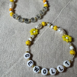 Può includere: Due braccialetti di perline. Un braccialetto presenta la parola "GEWOLLT" scritta con perline di lettere bianche. I braccialetti hanno perline gialle, bianche e arancioni, con alcuni accenti di perle. Un braccialetto ha perline a forma di fiore giallo.