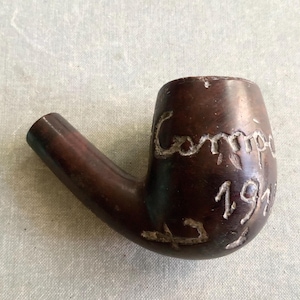 May include: A dark brown wooden tobacco pipe featuring a smooth, curved bowl and a short stem. The exterior of the bowl is hand-carved with the text "Compiègne" and the year "1916" etched into the surface, showing visible wear and texture.