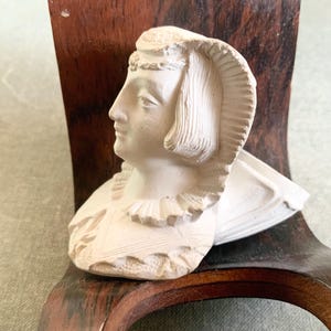 Puede incluir: Un busto de yeso blanco que representa el perfil de una persona con un tocado detallado, un cuello con volantes y ropa texturizada. La escultura presenta tallas intrincadas en el cabello, la corona y la prenda, mostrando un acabado mate.