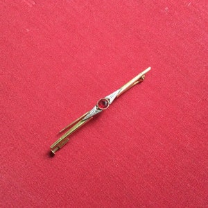 Könnte beinhalten: Eine goldfarbene Brosche mit einem roten Edelstein in der Mitte. Die Brosche ist in einer geometrischen Form mit einem spitzen Ende gestaltet.