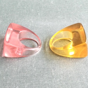 May include: Two transparent resin rings. One is a light pink color, and the other is a golden yellow. Both rings have a triangular shape with a rounded inner circle for the finger. The rings are displayed on a gray surface.