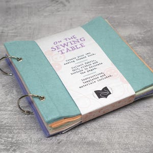 May include: A sewing-themed book with a light blue cover and silver binder rings. The book is wrapped with a white band that reads "ON THE SEWING TABLE" and includes instructions and templates. The pages are various colours.