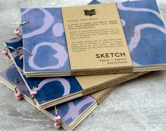 May include: A stack of three horizontal sketchbooks with blue and light purple abstract patterned covers. Each sketchbook is bound with metal rings featuring small pink beads. A tan paper band wraps around the center with printed text.