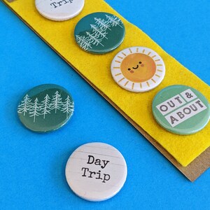 Peut inclure: Quatre badges à épinglette sur un fond jaune. Les badges représentent un soleil avec un visage souriant, une scène de forêt et le texte "Day Trip" répété deux fois. Le texte "OUT & ABOUT" est sur le quatrième badge.