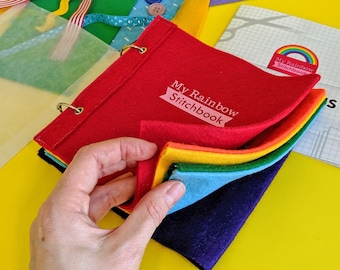 Könnte beinhalten: Ein rotes Filzbuch mit dem Titel "My Rainbow Stitchbook" mit Regenbogenfarbenen Filzseiten. Das Buch ist geöffnet und zeigt die Schichten. Weitere Filzteile und Verzierungen sind sichtbar, darunter Knöpfe und Bänder.