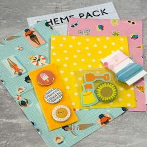 May include: A summer-themed craft kit featuring yellow polka dot fabric, blue and white striped fabric, pink fabric with beach-themed illustrations, and a sheet of stickers with a beach shovel, bucket, and sunflower. The kit also includes three felt buttons with a popsicle, a sun, and the word "SUMMER" on them.