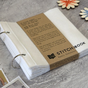 May include: A white fabric sketchbook with a brown paper cover. The cover has the text "STITCH IBOOK Fabric Sketchbook" and details about the materials used. The sketchbook is bound with silver rings.